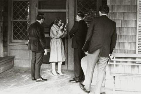 "Little" Edie Beale used a magnifying glass to read a search order from the Suffolk Health Department in October, 1971, as officials watched on the porch of Grey Gardens, where she lived with her mother, Edith Bouvier Beale. The department had responded to reports that the two women were harboring diseased cats and that the rundown house's furnace was unsafe. "Miss Beale called the inspection 'a raid,' engineered by henchmen of 'a mean, nasty Republican Town,'" in a page-one story.